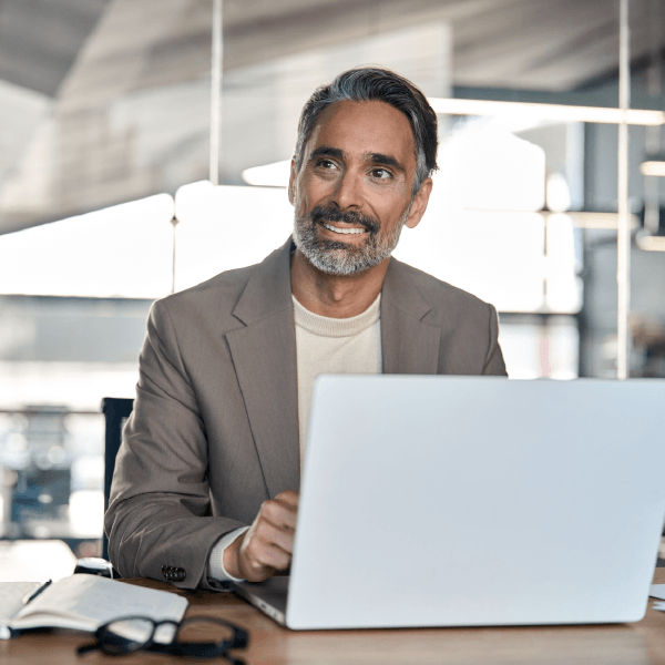 Person in office setting looking up smiling, with glasses, notepad and laptop.